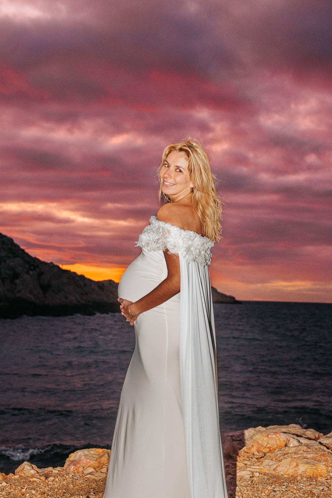 Future maman en robe blanche face à la mer sous un ciel rose de coucher du soleil