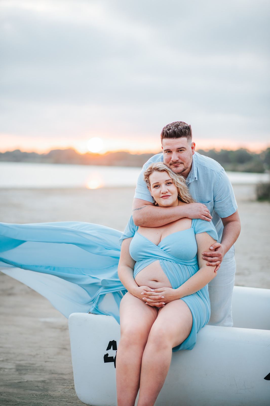 Future maman en robe bleue fluide qui vole au vent, assise au bord de l'eau