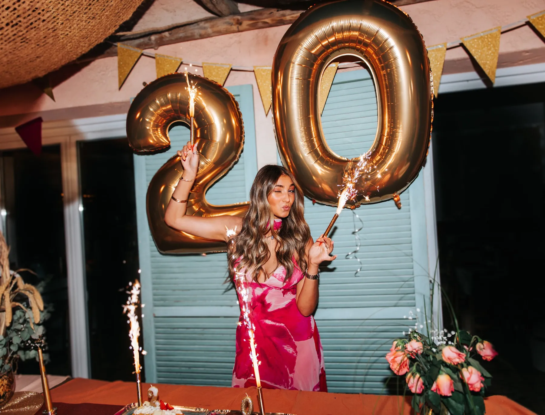 Anniversaire des 20 ans : jeune femme en robe rose qui fait un clin d'œil, bougies étincelles et ballons dorés