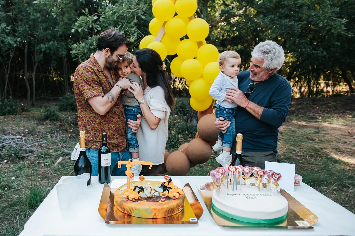 Anniversaire en famille dans un jardin : câlins autour de deux gâteaux, devant une arche de ballons jaunes