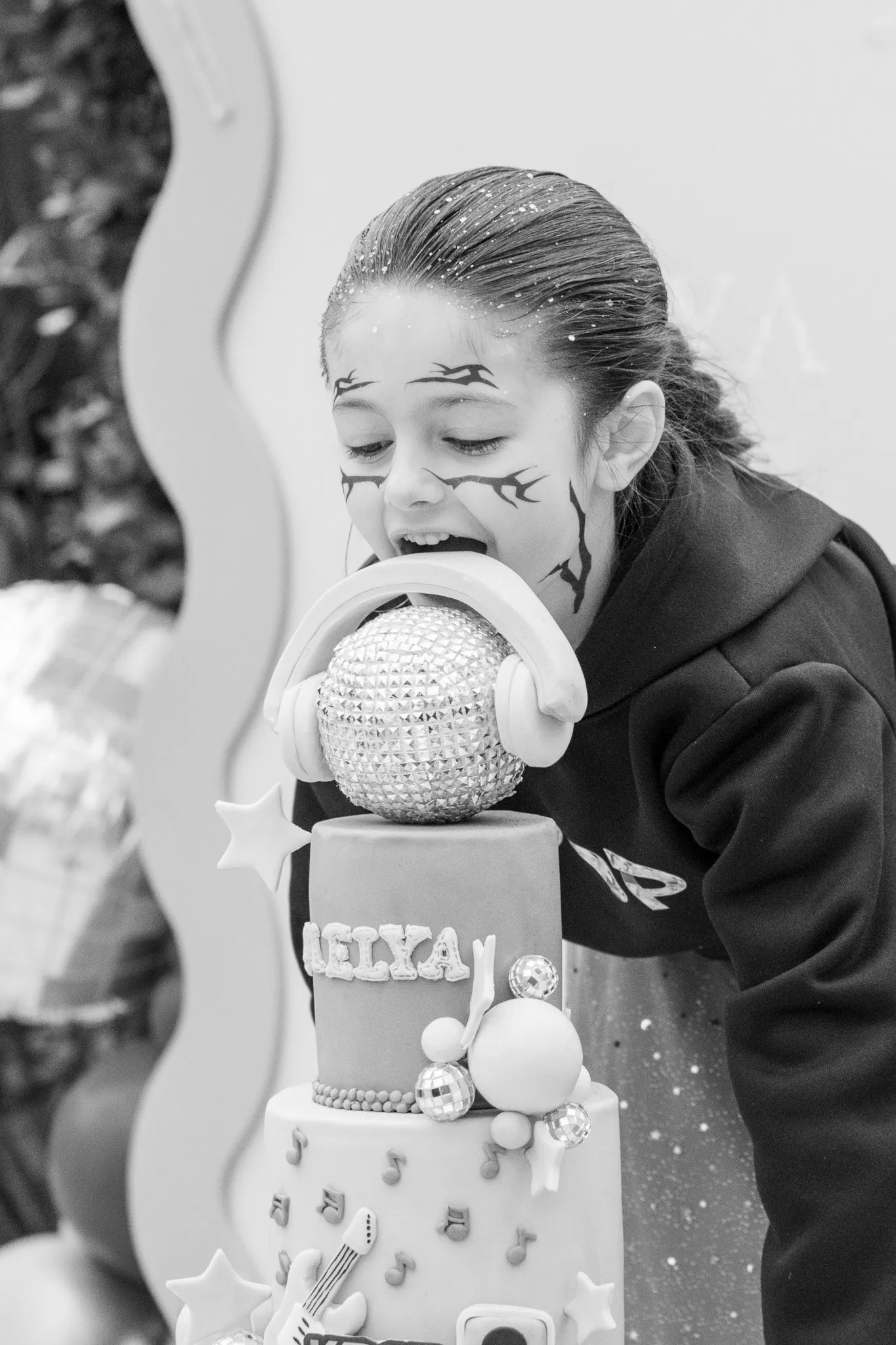 Fête d'anniversaire : fillette maquillée qui fait mine de croquer la boule disco de son gâteau, en noir et blanc