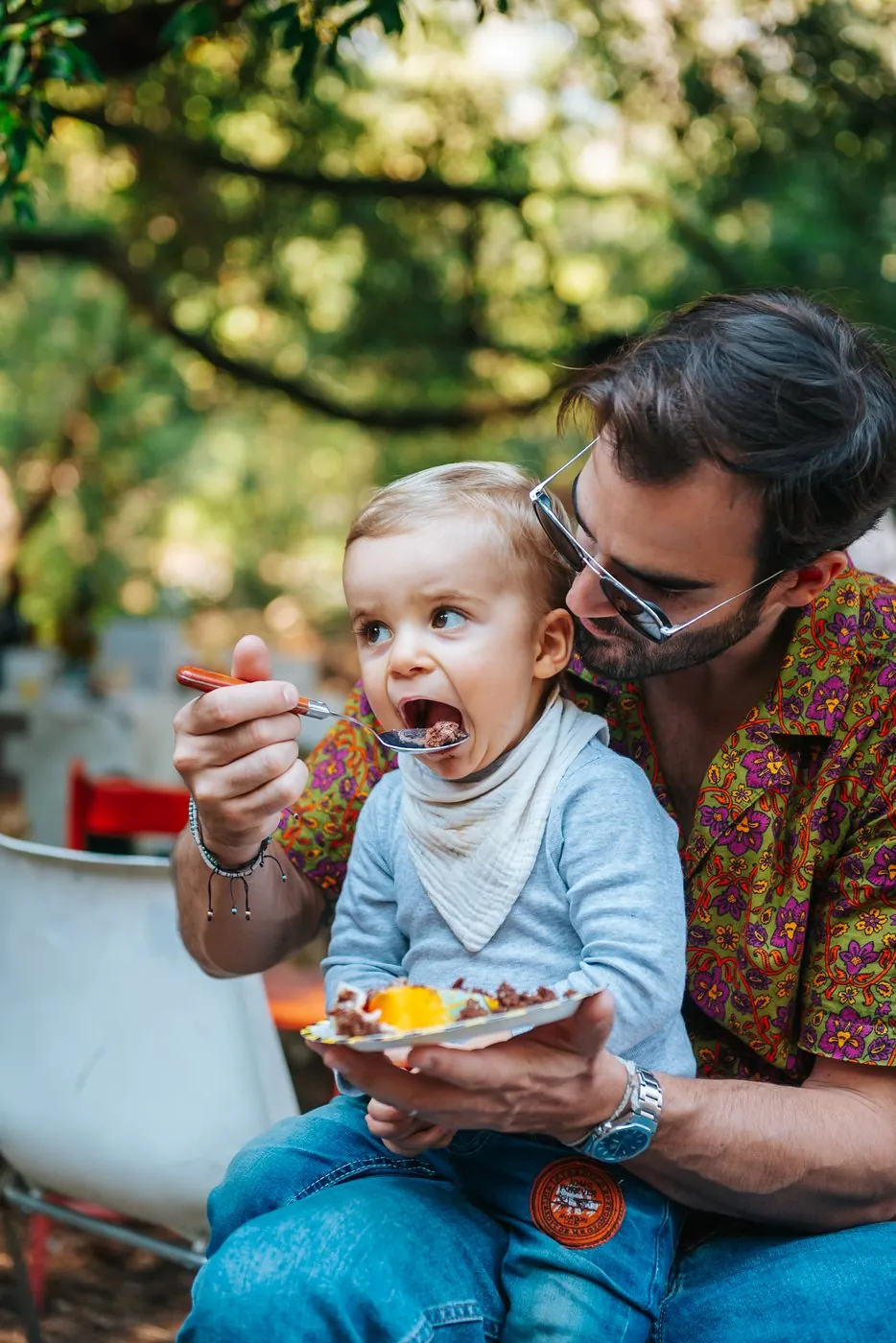 Fête d'anniversaire en plein air : un papa donne une cuillère de gâteau à son petit garçon, bouche grande ouverte