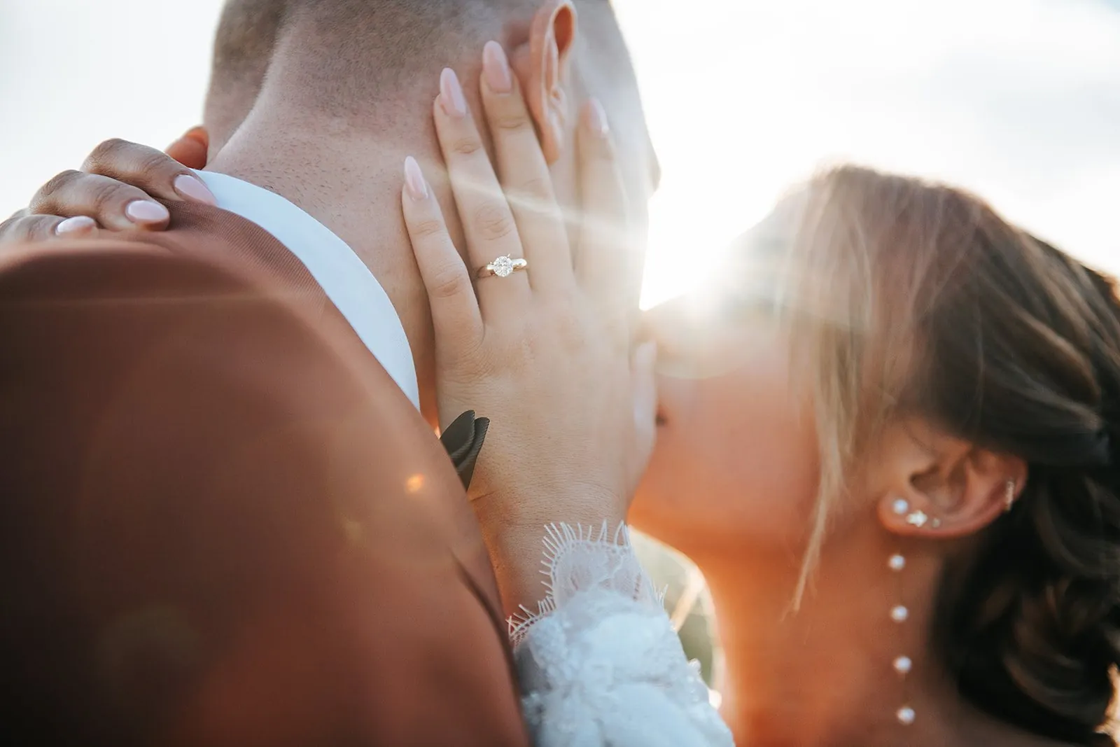 Gros plan sur la bague de fiançailles de la mariée, sa main sur la nuque du marié, dans un rayon de soleil