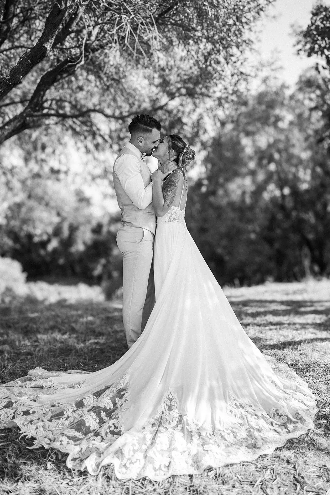 Mariage en noir et blanc : baiser des mariés sous les arbres, la longue traîne en dentelle déployée