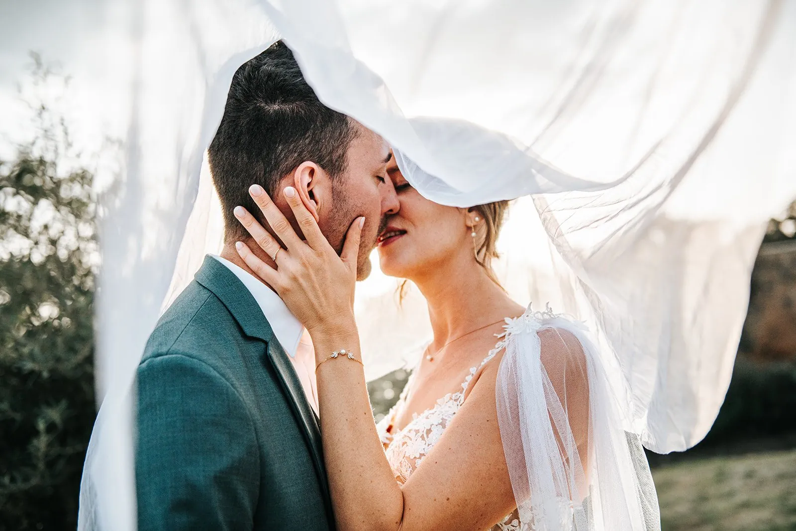 Mariage : baiser des mariés sous un voile blanc soulevé par le vent, la main de la mariée posée sur sa joue