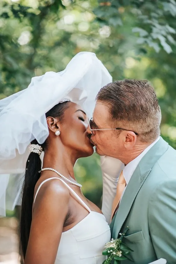 Mariage : baiser des mariés sous le voile soulevé par le vent, collier de perles et verdure en arrière-plan