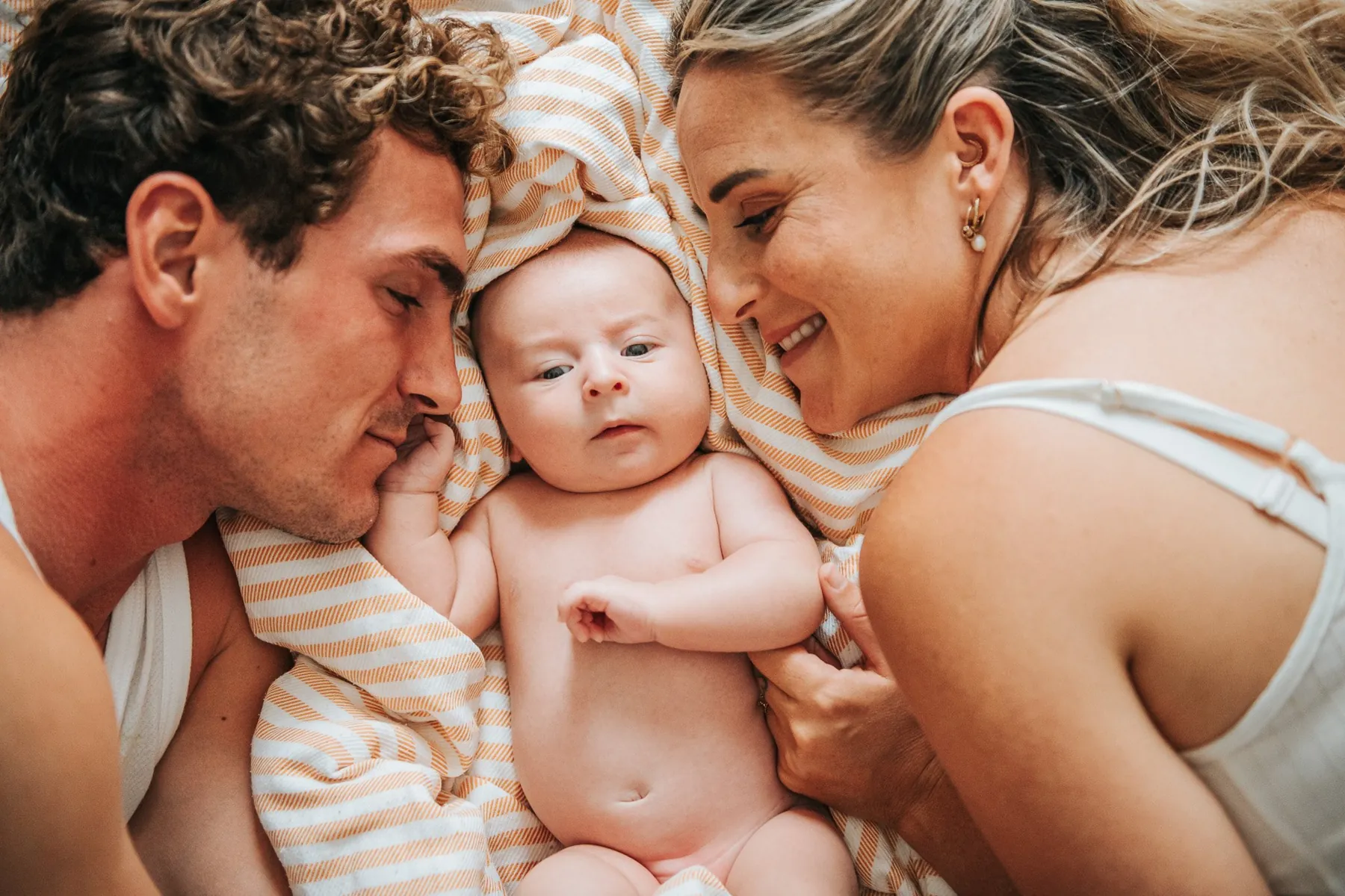 Baby lying on a striped cloth between smiling parents, faces close together, a tender moment at home