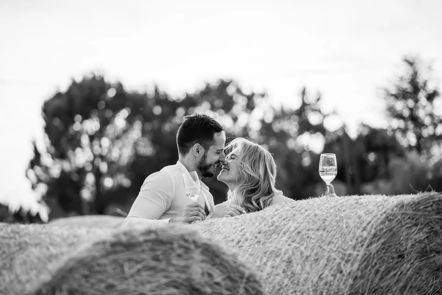 Couple complice nez contre nez derrière des bottes de foin, verre de vin à la main, en noir et blanc
