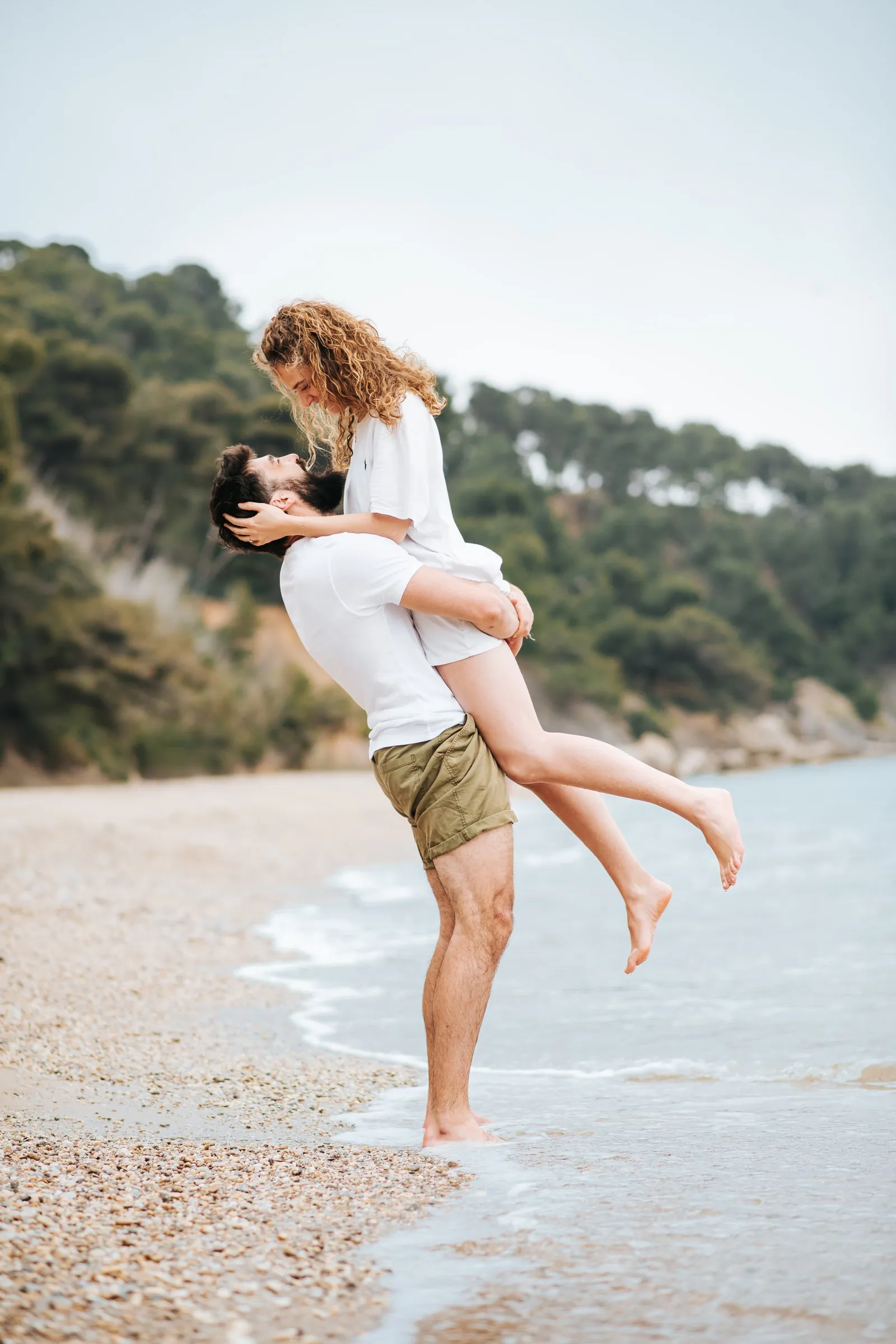 Sur une plage de galets, il soulève sa compagne dans ses bras, pieds dans l'eau, face aux pins en bord de mer