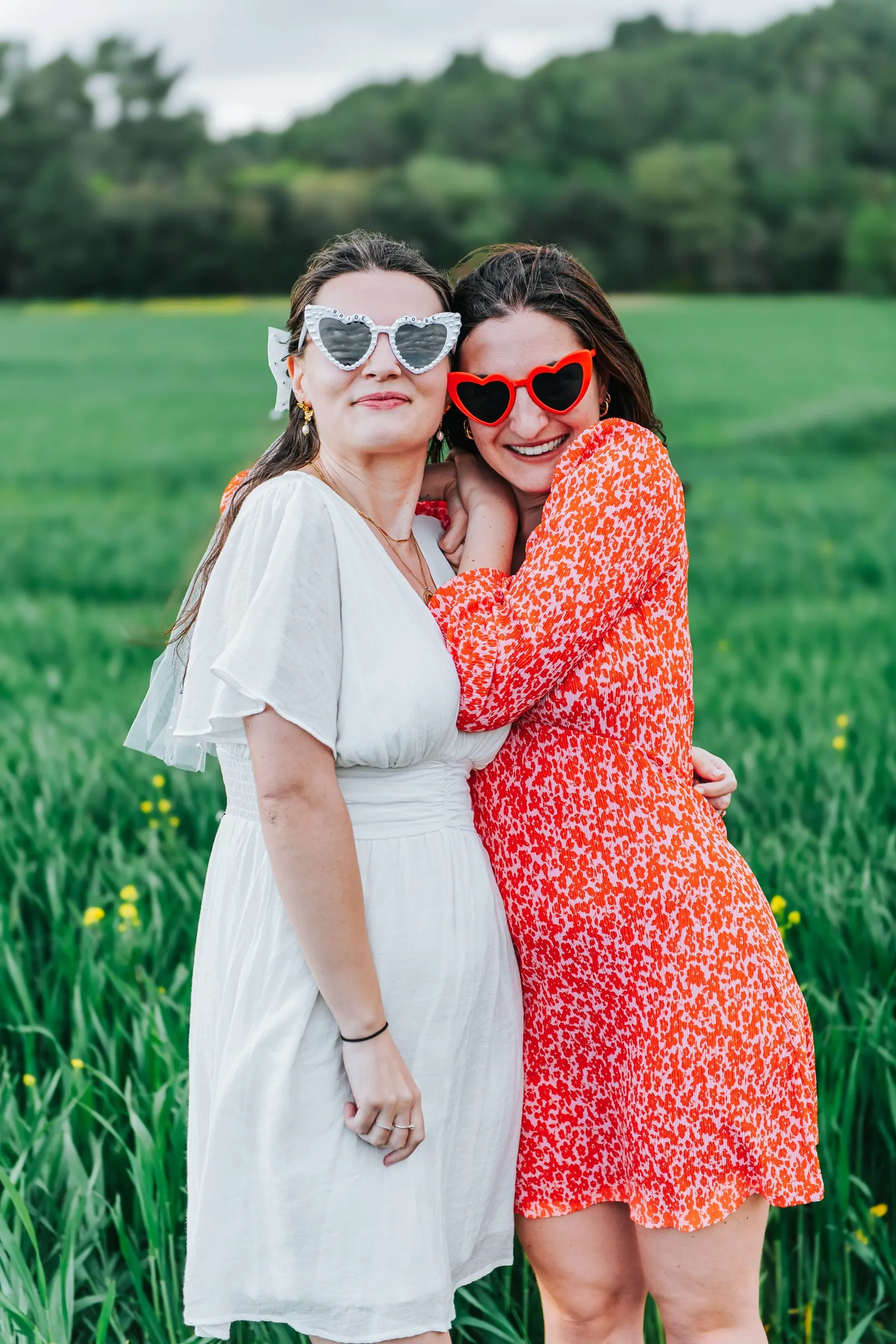 Enterrement de vie de jeune fille : la future mariée et son amie enlacées, lunettes cœur sur le nez, dans un pré
