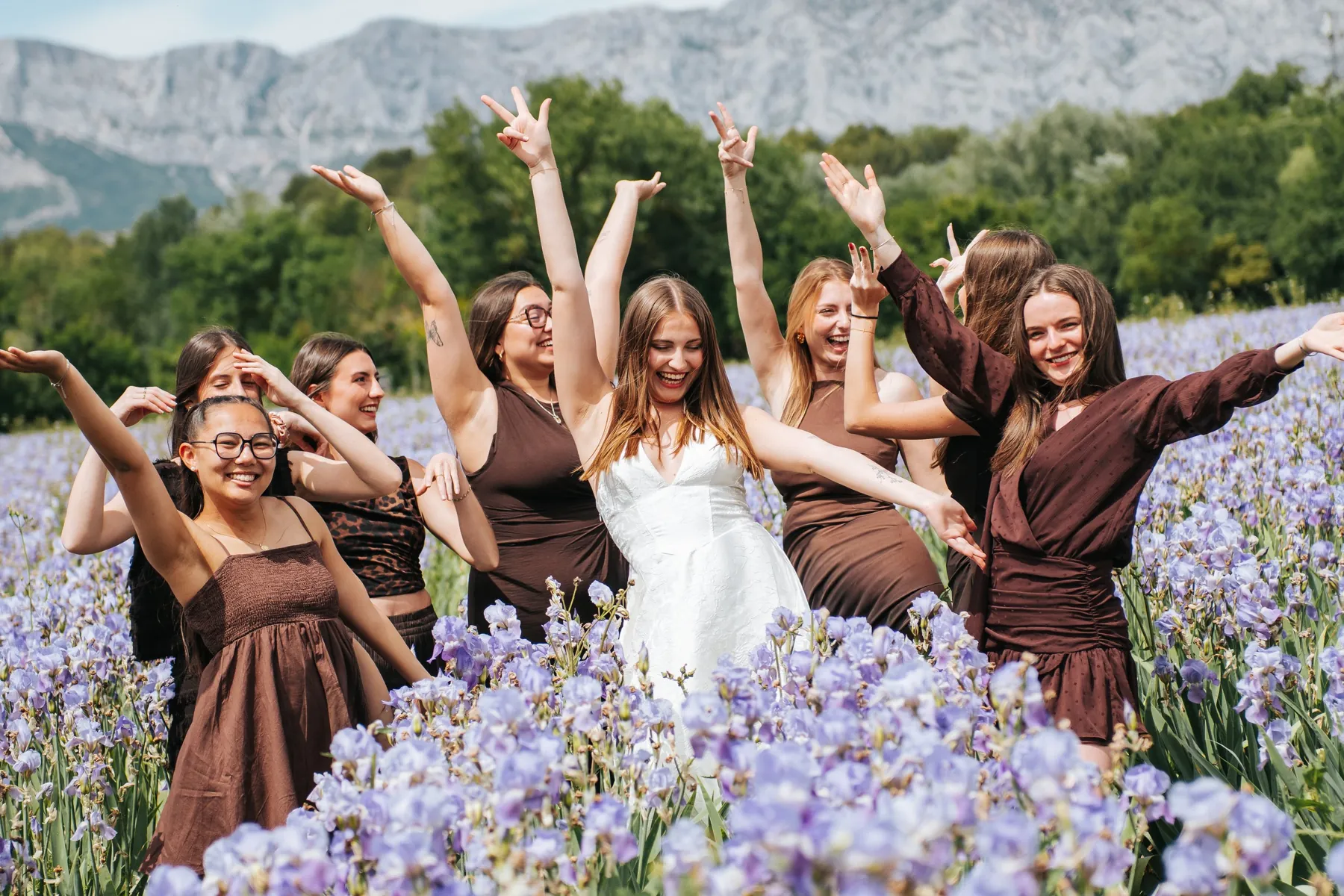 EVJF dans un champ d'iris : la future mariée en blanc et ses amies en robes marron lèvent les bras en riant, près d'Aix-en-Provence