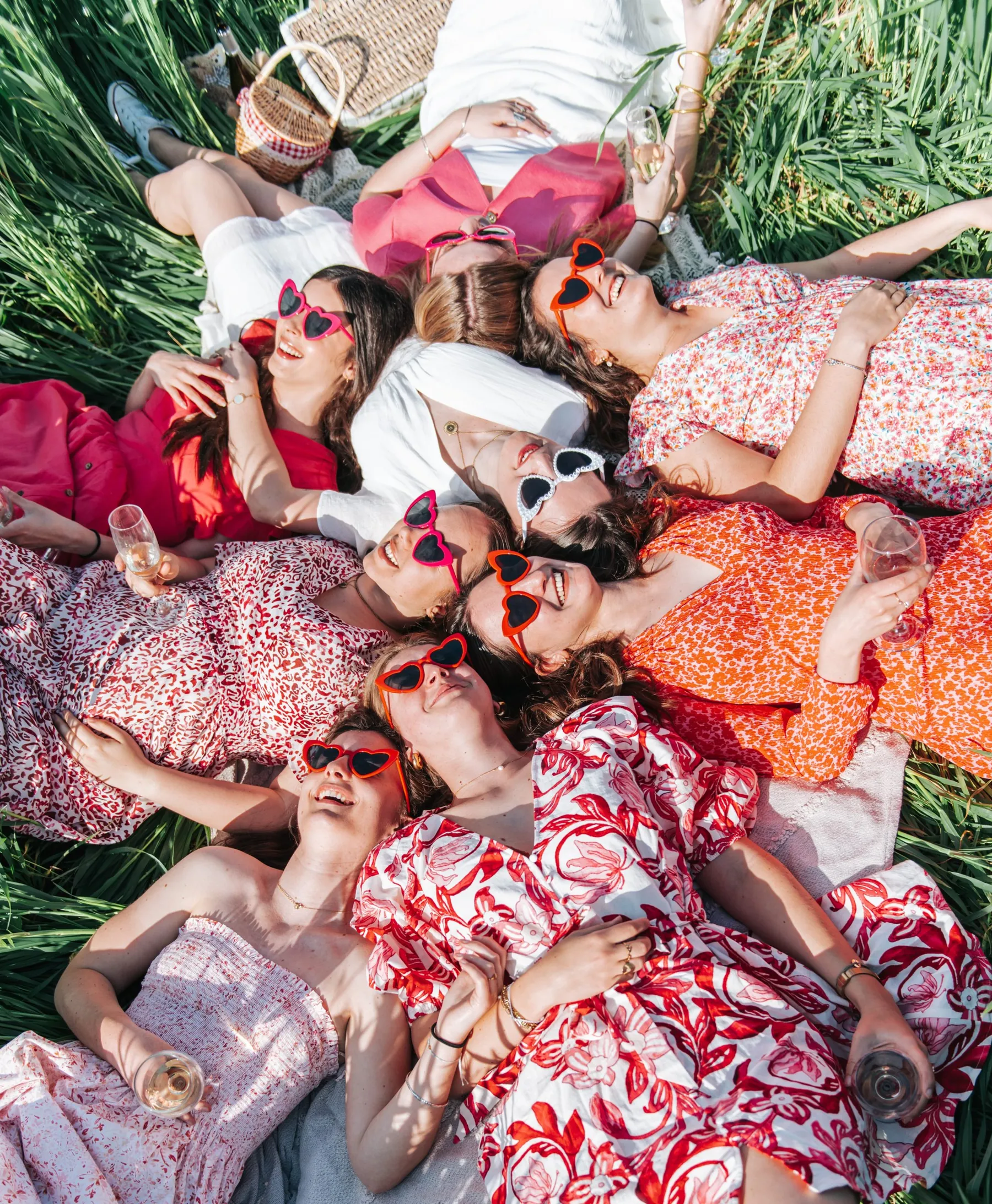 EVJF pique-nique : copines allongées en étoile dans l'herbe, lunettes cœur et coupe à la main, éclats de rire
