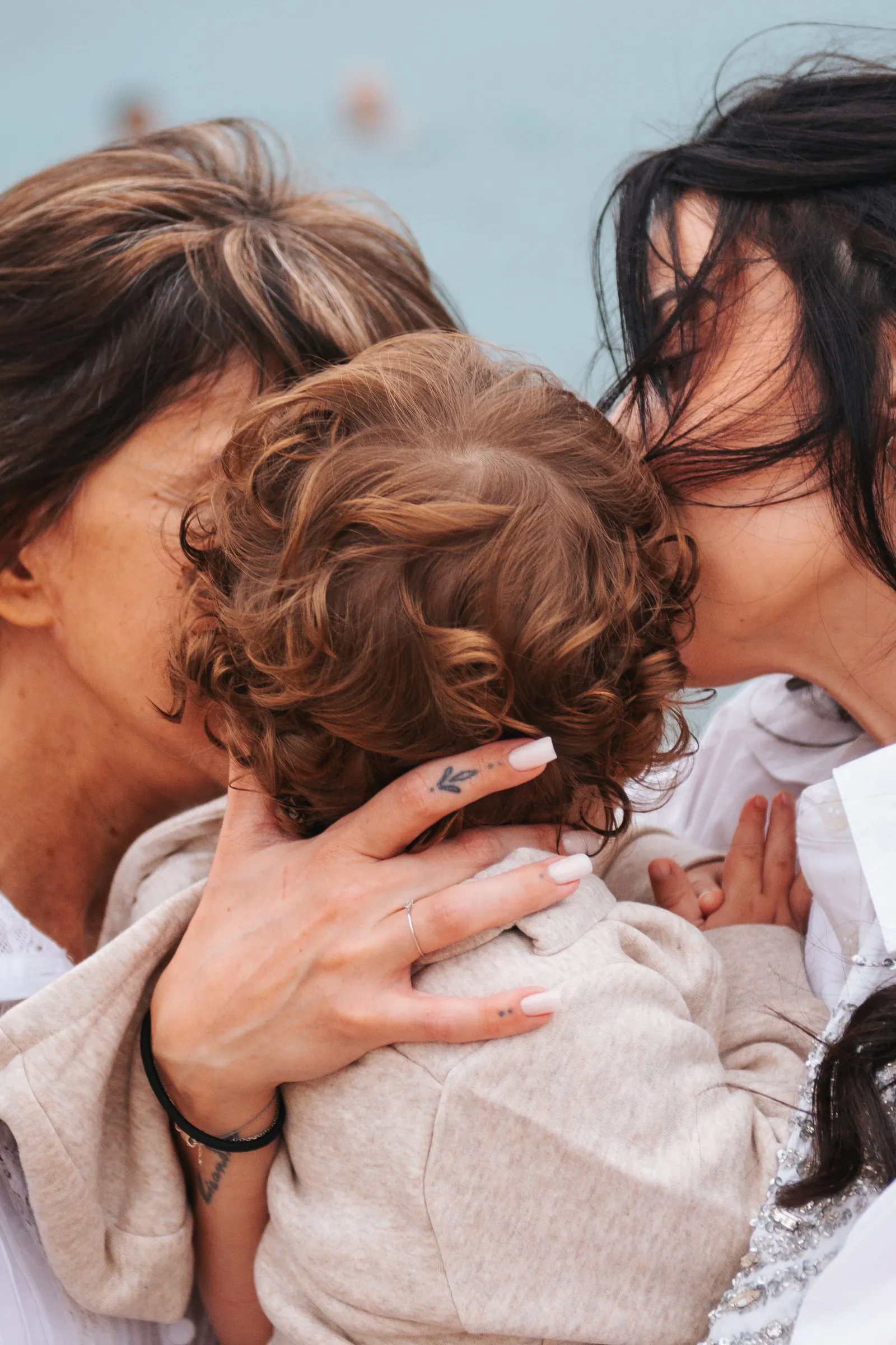 Two mums kissing their baby's curly head by the sea, a close-up full of tenderness