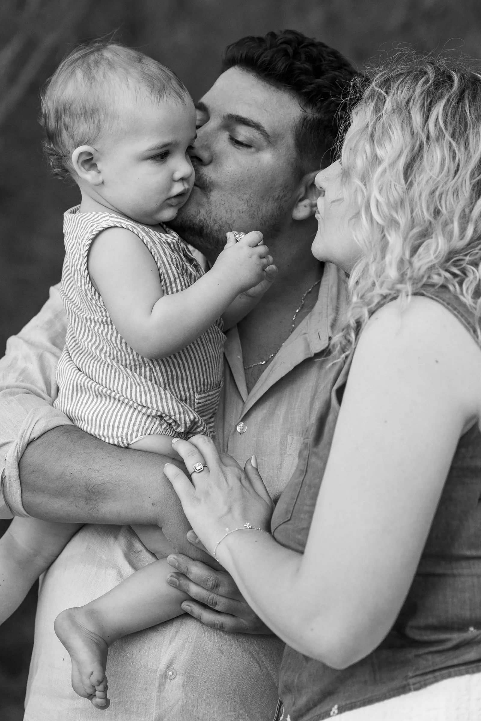 A dad kissing the cheek of the baby in his arms, mum close by, a tender family portrait in black and white
