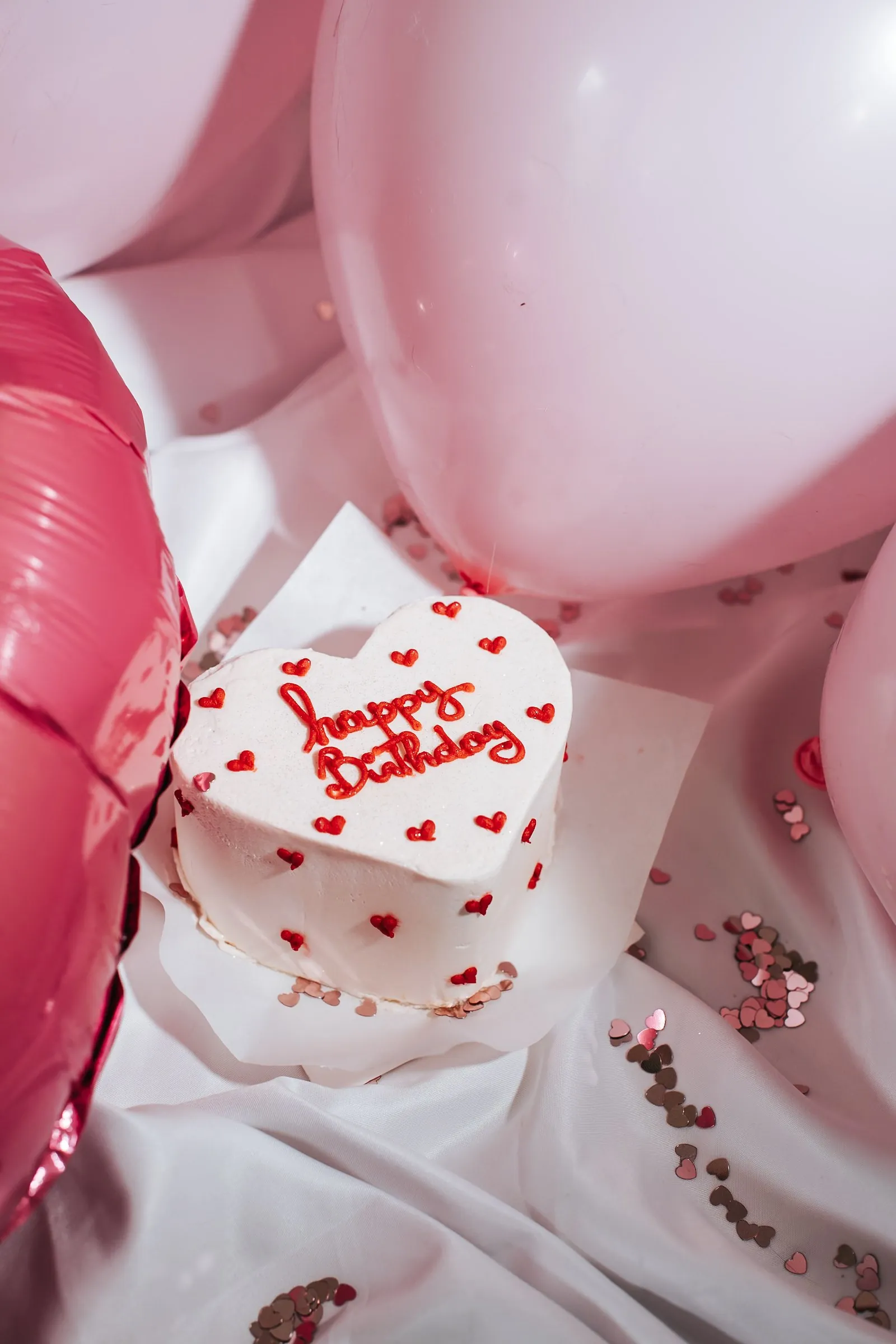 Gâteau d'anniversaire en forme de cœur « Happy Birthday » entouré de ballons roses et de confettis cœurs