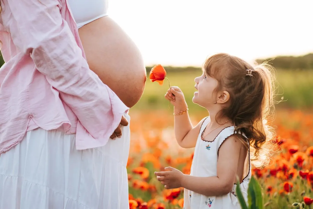 Séance grossesse dans un champ de coquelicots : petite fille qui tend une fleur vers le ventre rond de sa maman