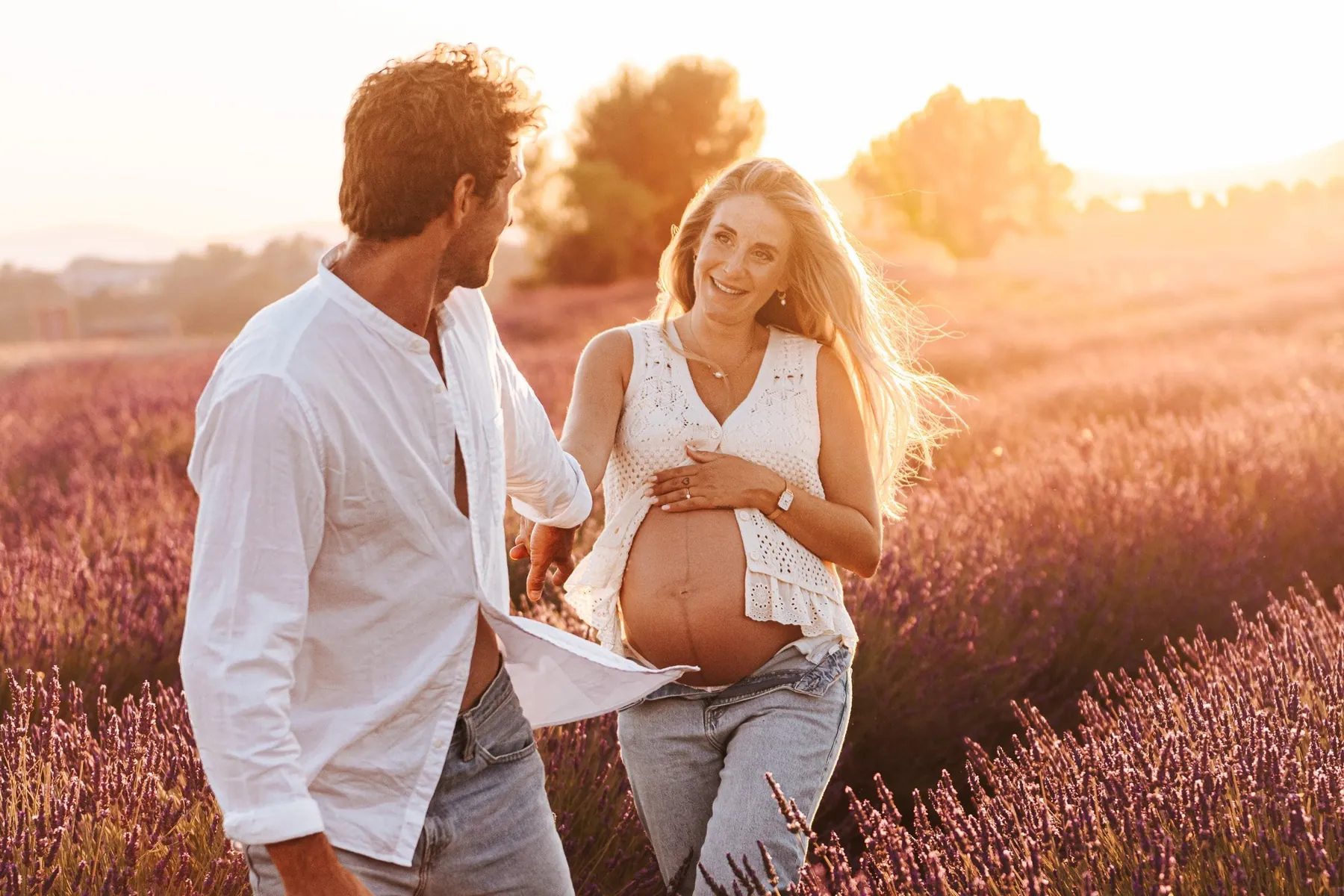 Couple complice qui marche dans un champ de lavande au coucher du soleil, la future maman tenant son ventre nu