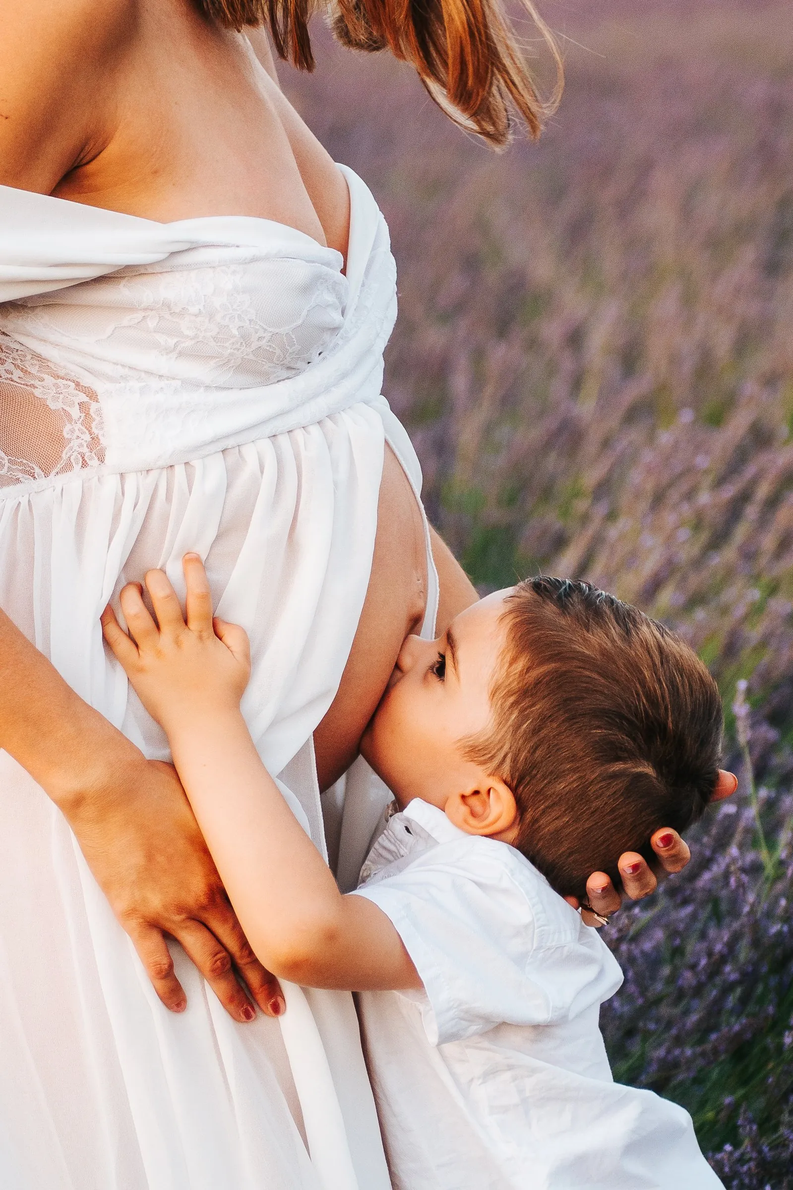 Séance grossesse dans la lavande : petit garçon qui embrasse le ventre rond de sa maman en robe blanche