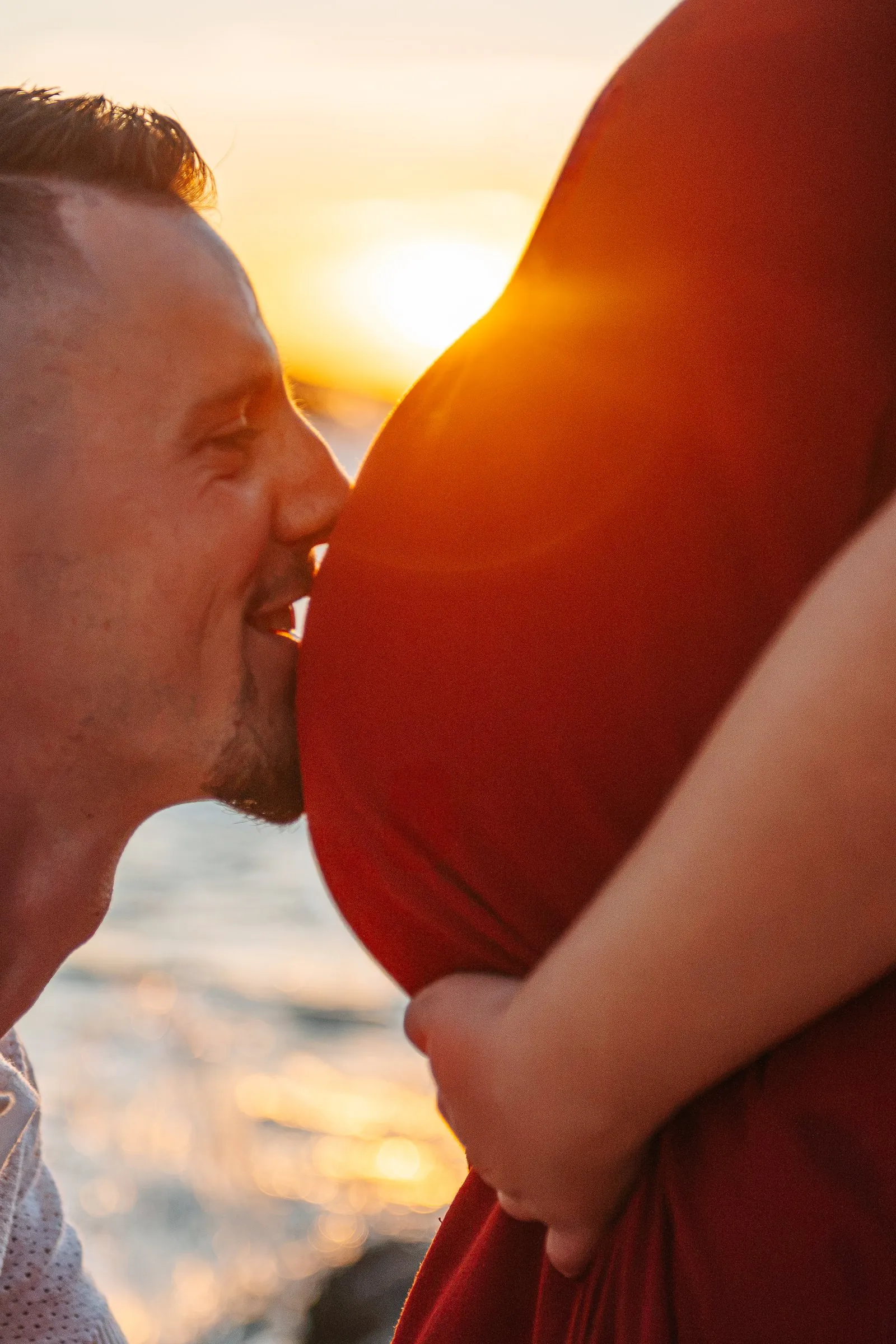 Close-up of a dad-to-be kissing his partner's bump in a red dress, with the sun setting over the sea