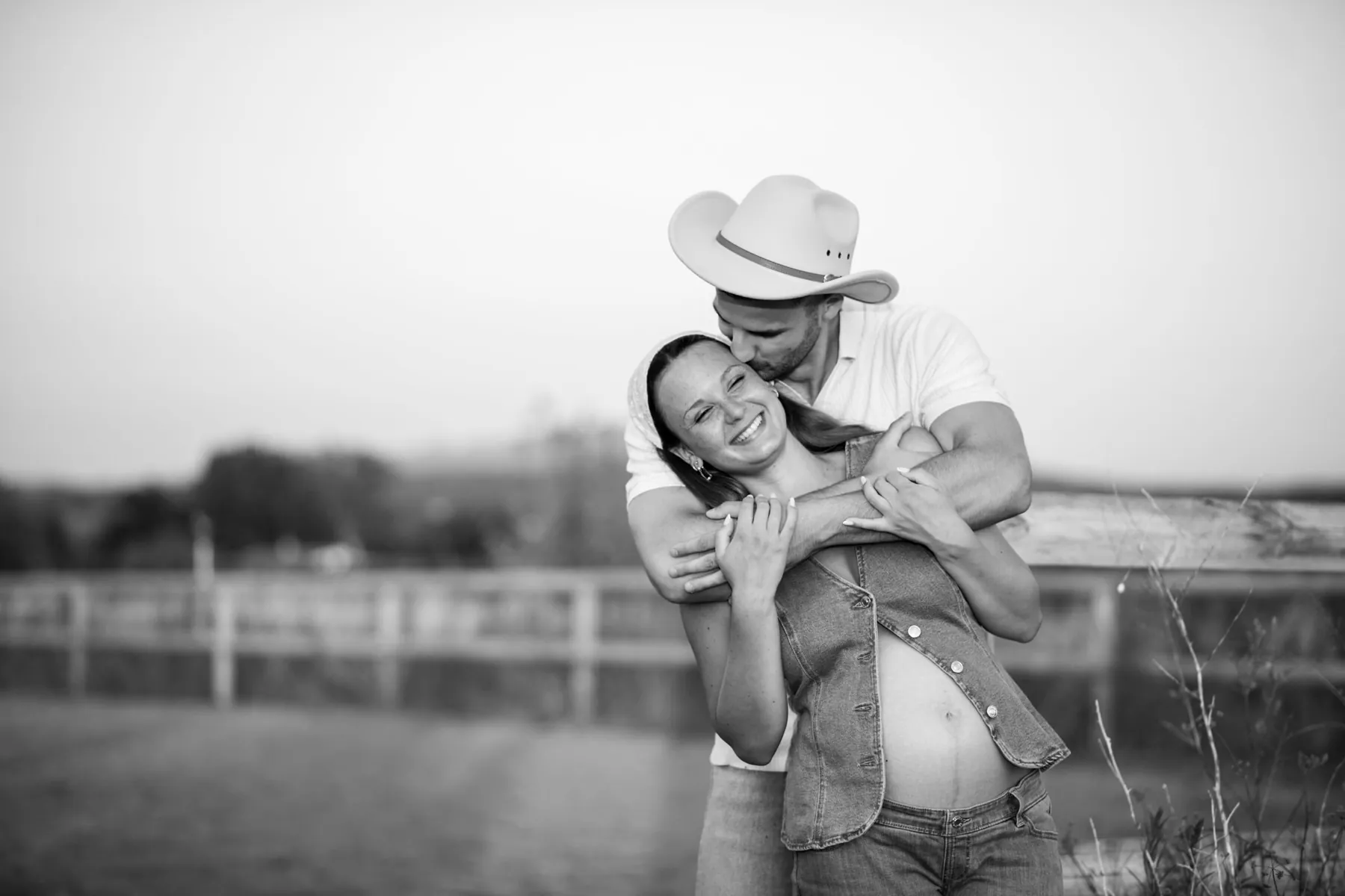 Future maman en gilet en jean qui éclate de rire dans les bras de son compagnon au chapeau de cowboy, en noir et blanc, près d'Aix-en-Provence