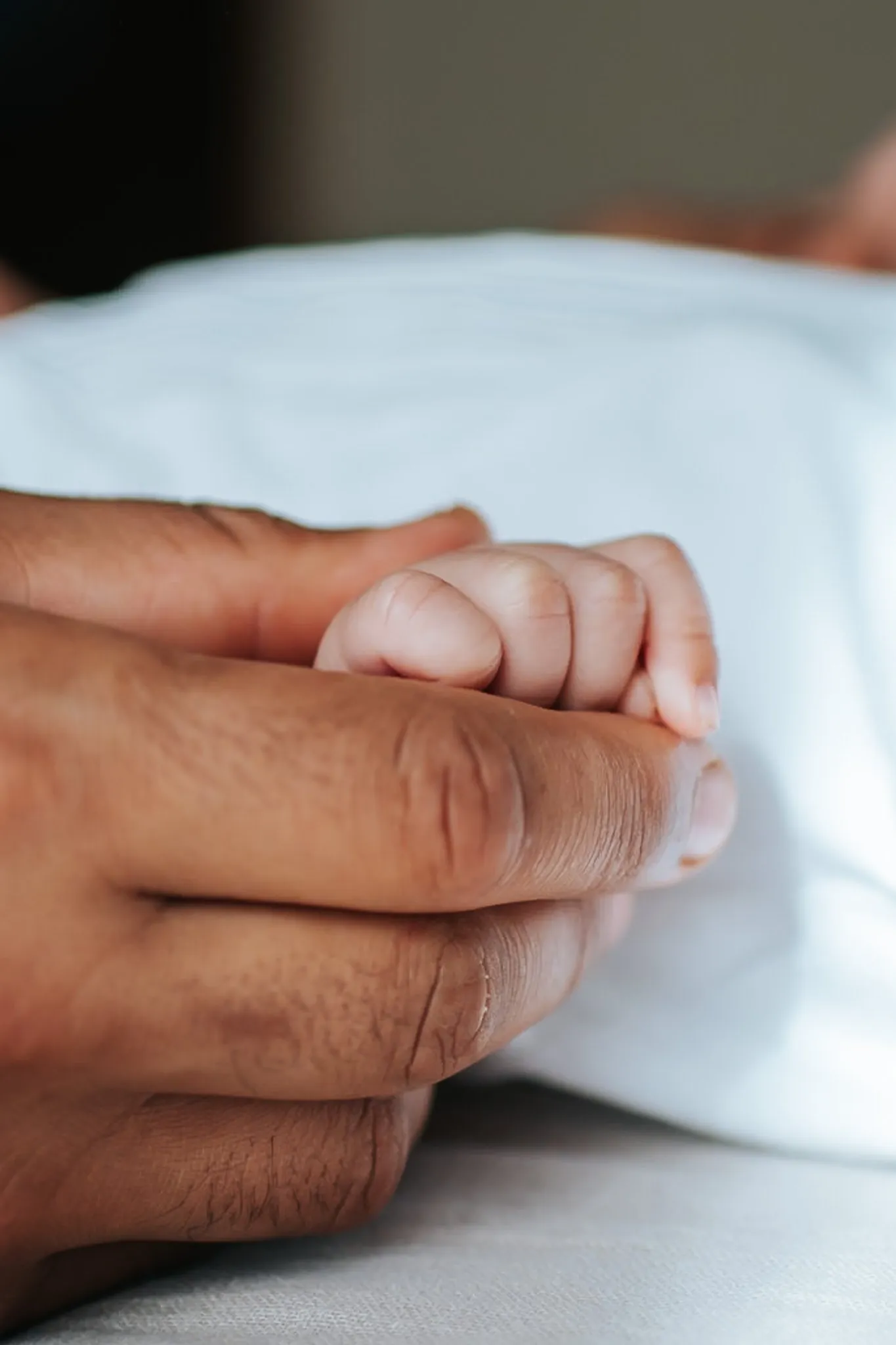 Minuscule poing de bébé serré dans la main de son papa, sur un drap blanc, en lumière douce et naturelle