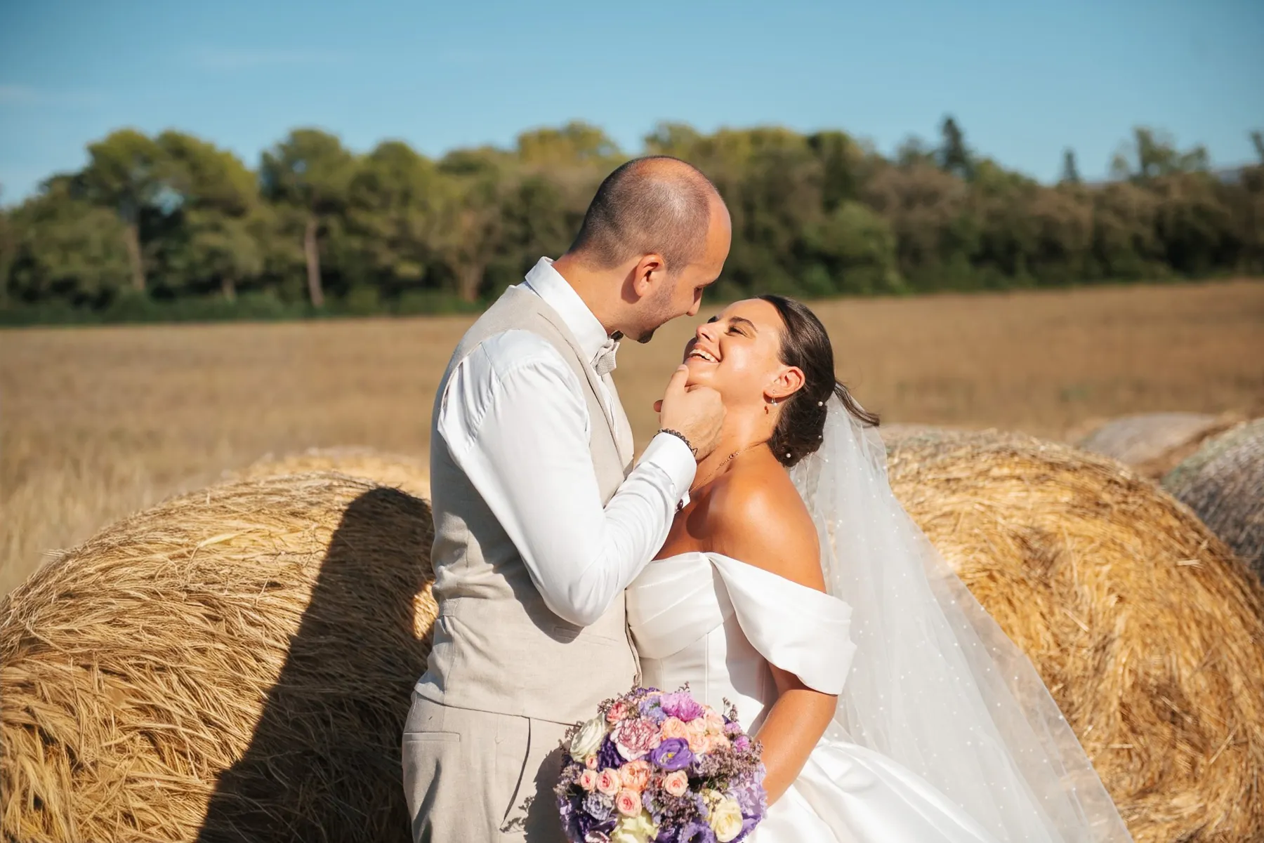 Mariage champêtre : le marié caresse le visage de la mariée rieuse, adossés à des bottes de paille au soleil