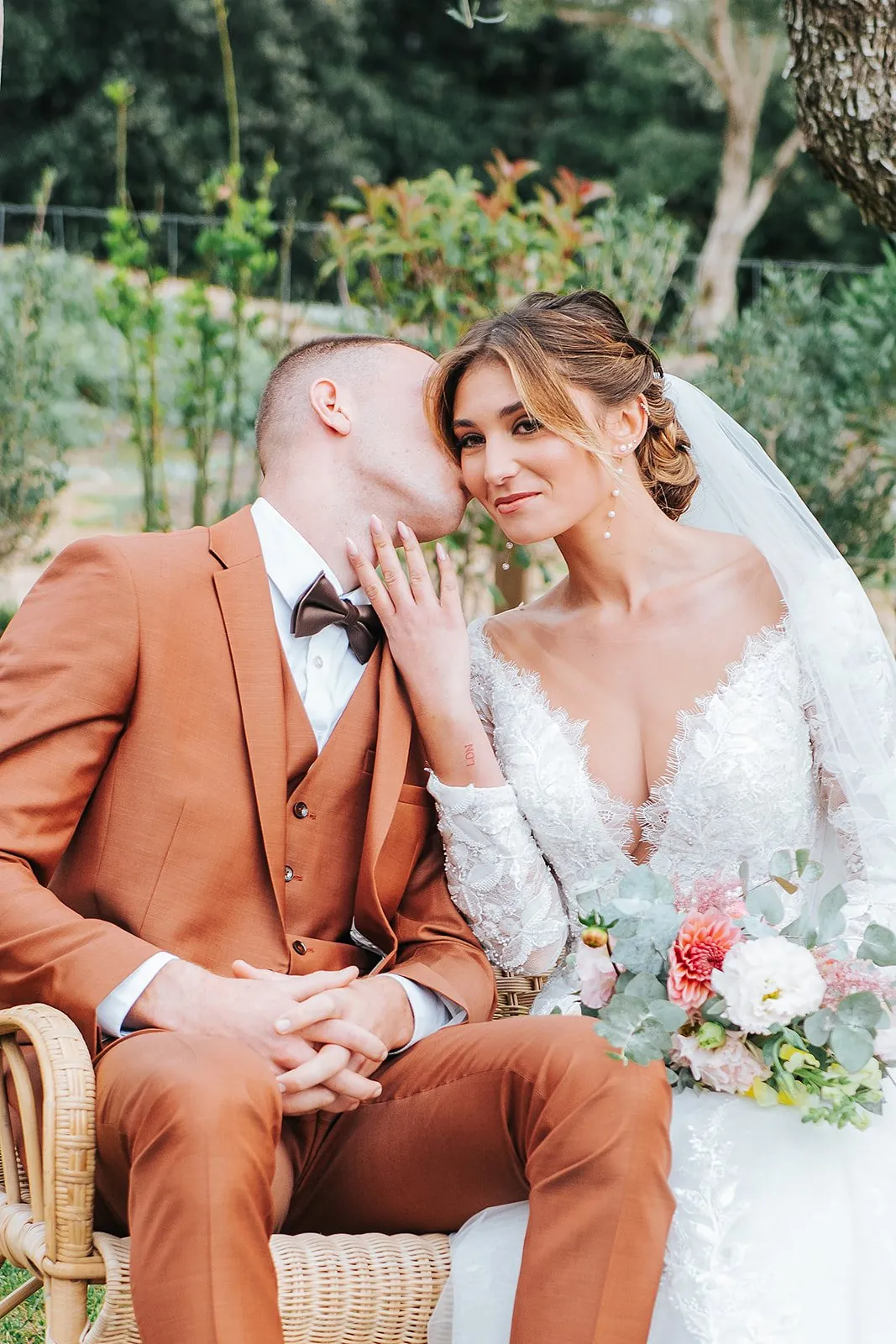 Mariage champêtre : le marié en costume terracotta embrasse la joue de la mariée, assis dans un fauteuil en rotin