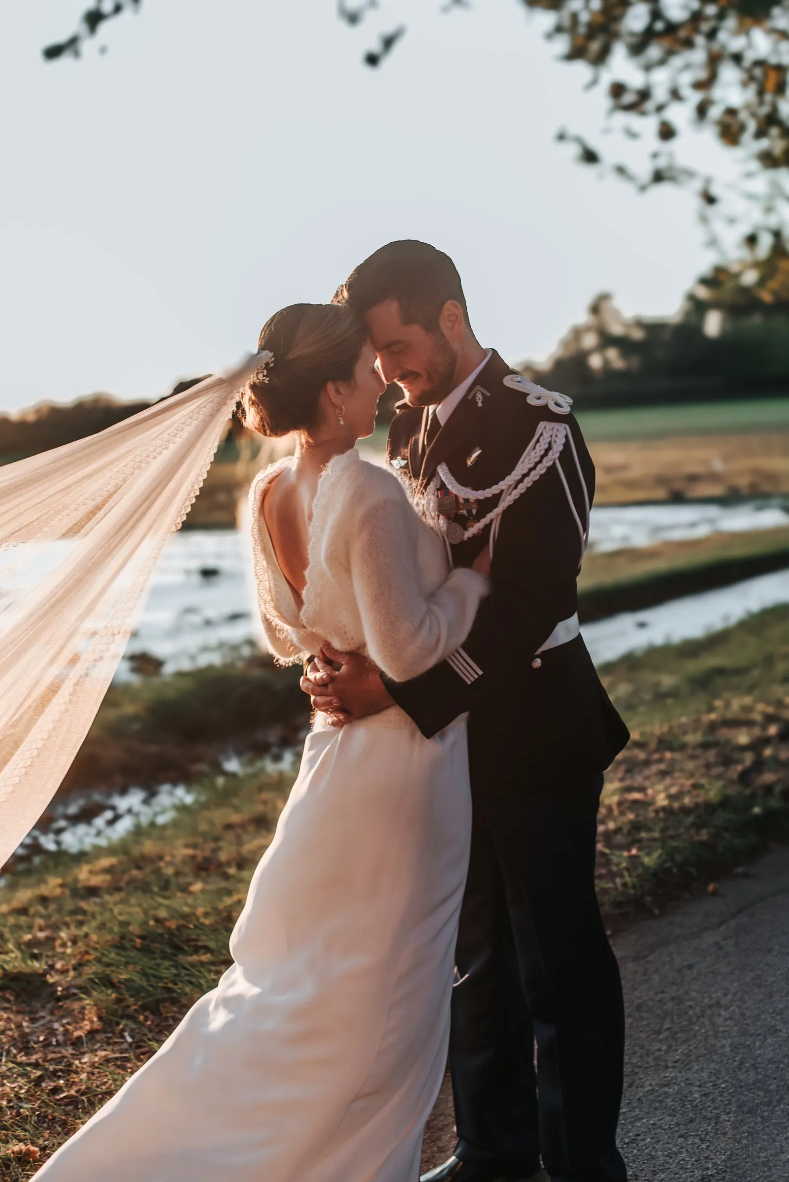 Mariage militaire : les mariés front contre front au soleil couchant, le long voile de la mariée porté par le vent
