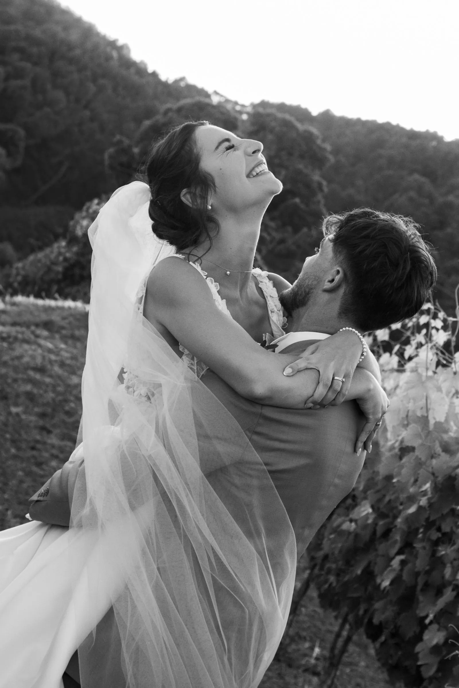 Le marié soulève la mariée qui éclate de rire, tête en arrière, au milieu des vignes, en noir et blanc