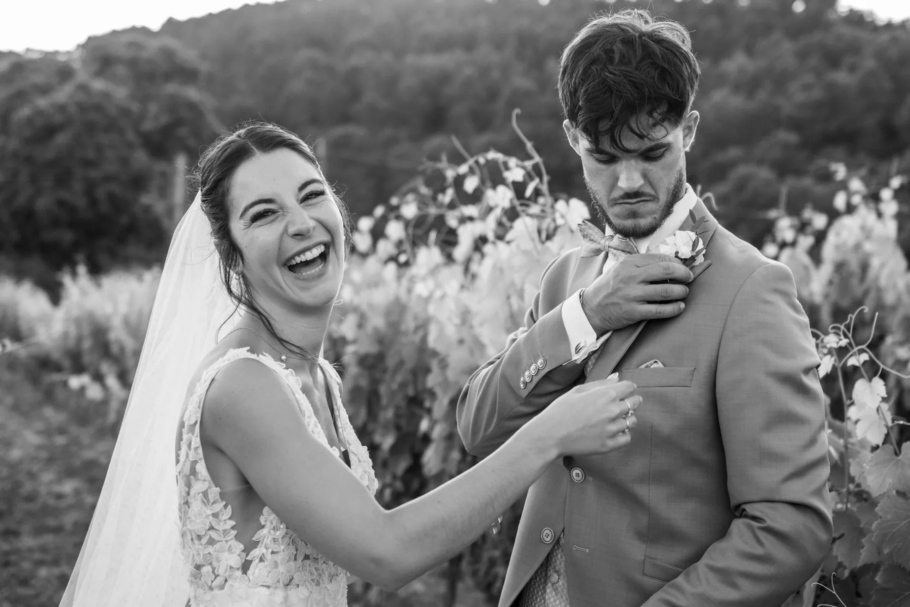 Mariage en noir et blanc dans les vignes : la mariée éclate de rire en ajustant la boutonnière du marié