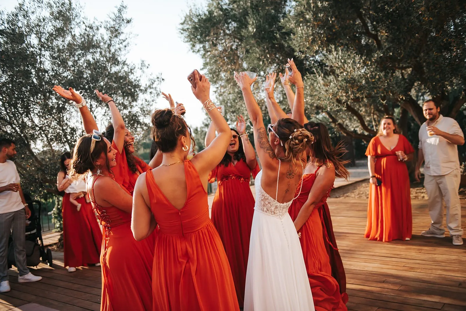 Mariage : la mariée danse bras levés avec ses demoiselles d'honneur en robes orange, sous les oliviers