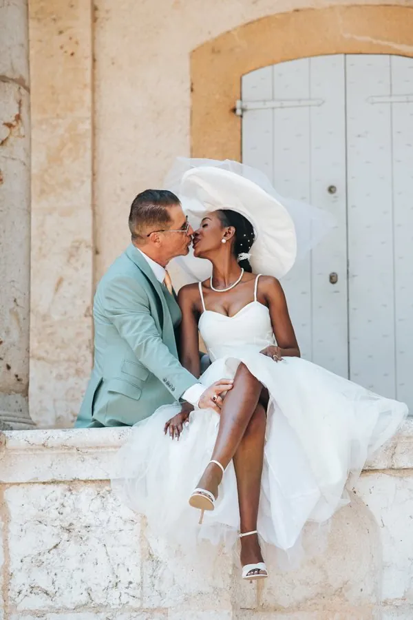 Mariée en robe courte en tulle assise sur un muret de pierre, embrassant le marié en costume vert d'eau