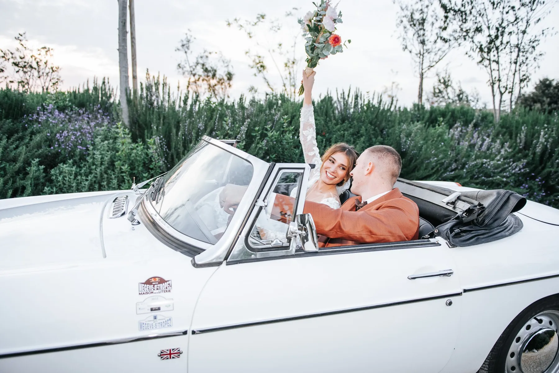 Les mariés partent en cabriolet blanc ancien, la mariée lève son bouquet