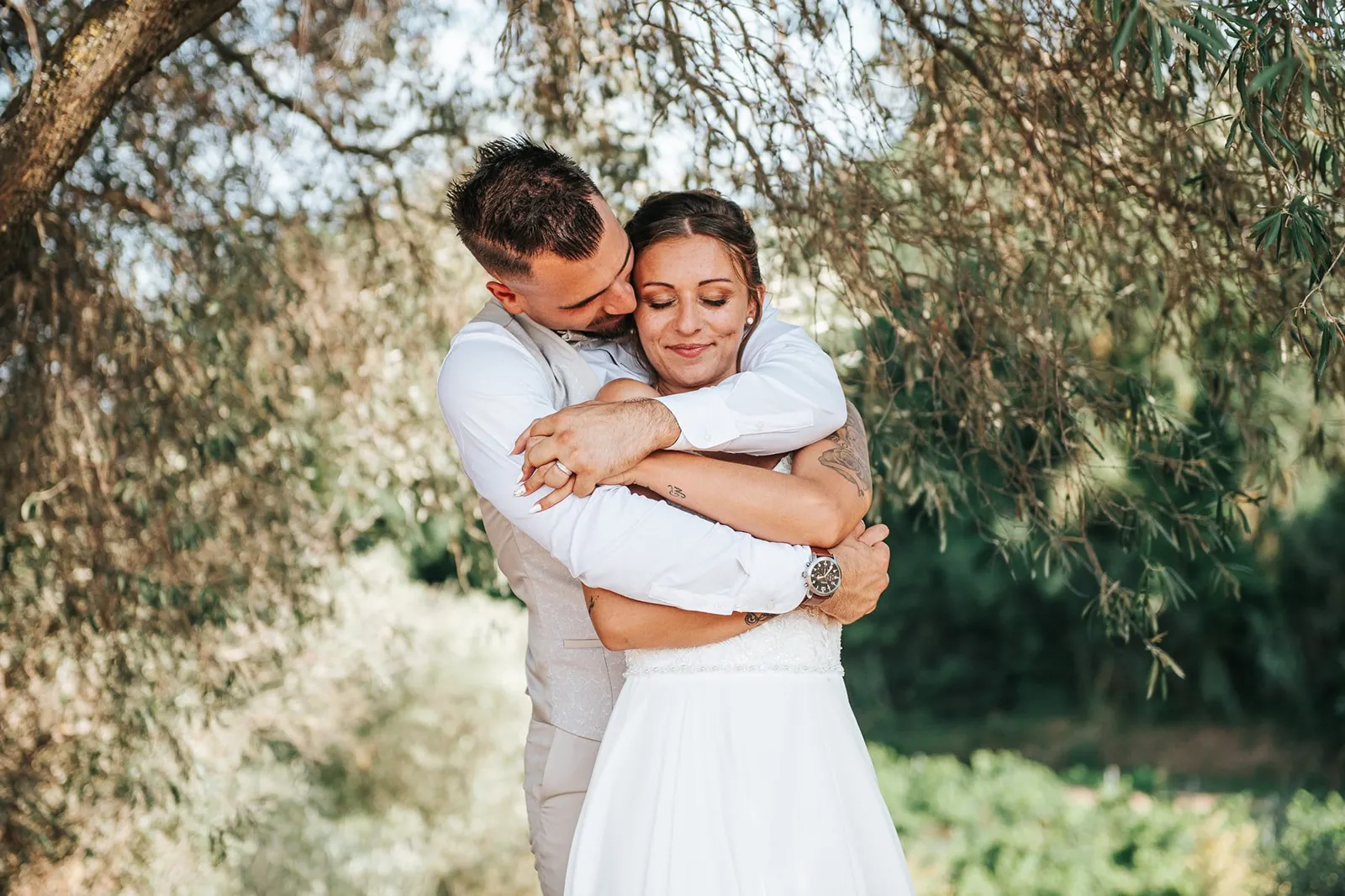 Le marié enlace tendrement la mariée par derrière, yeux fermés et sourire paisible, à l'ombre d'un olivier
