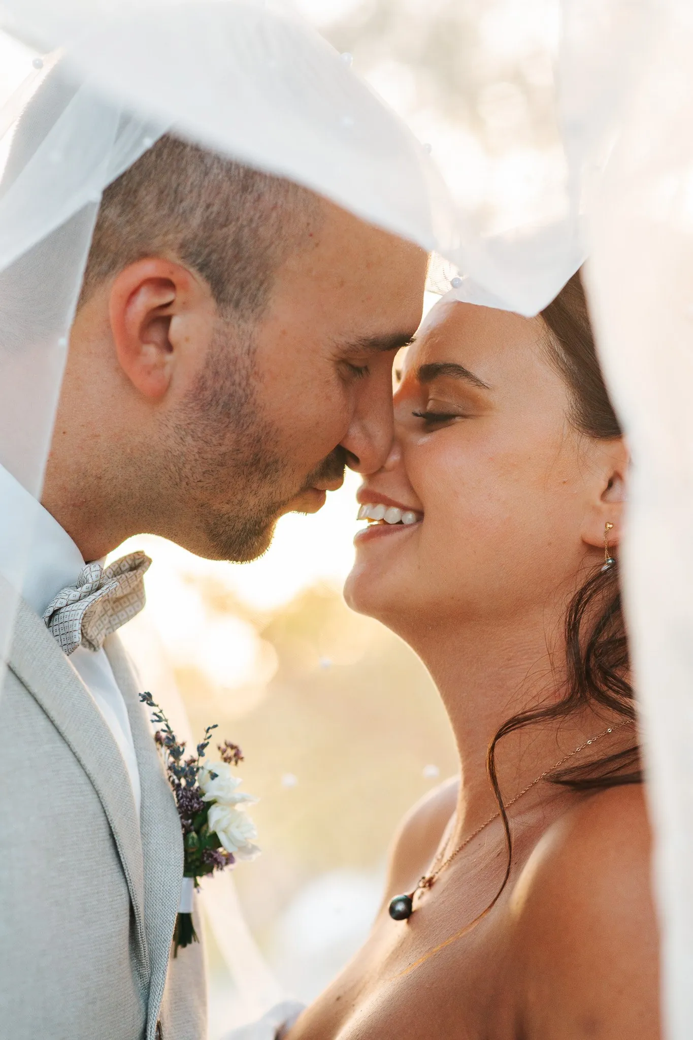 Wedding: the couple nose to nose beneath the veil, the bride laughing with eyes closed in golden light