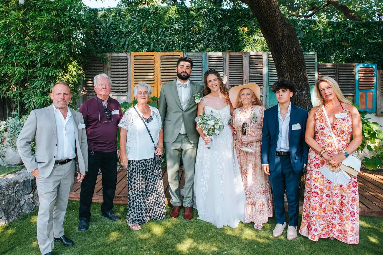Photo de groupe au mariage : les mariés entourés de leur famille dans un jardin ombragé aux volets colorés