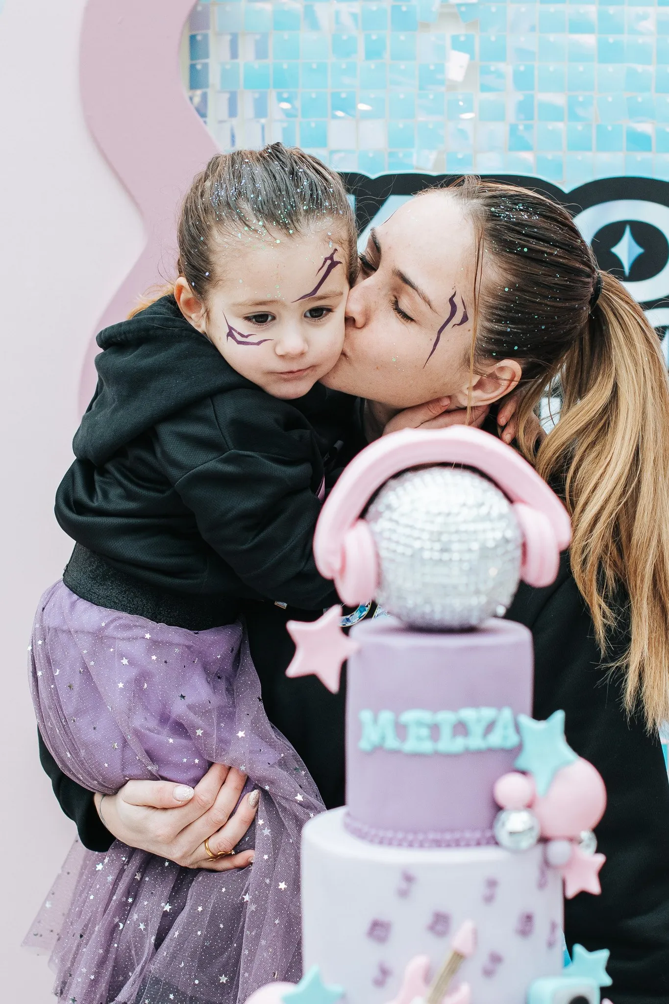 Anniversaire enfant : maman qui embrasse sa fille maquillée et pailletée devant un gâteau disco rose et violet