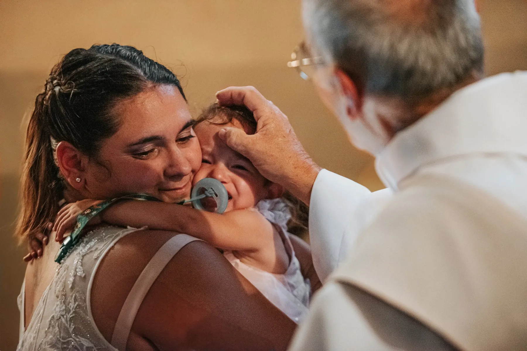 Baptême à l'église : le prêtre pose la main sur la tête d'une petite fille en pleurs, blottie contre sa maman