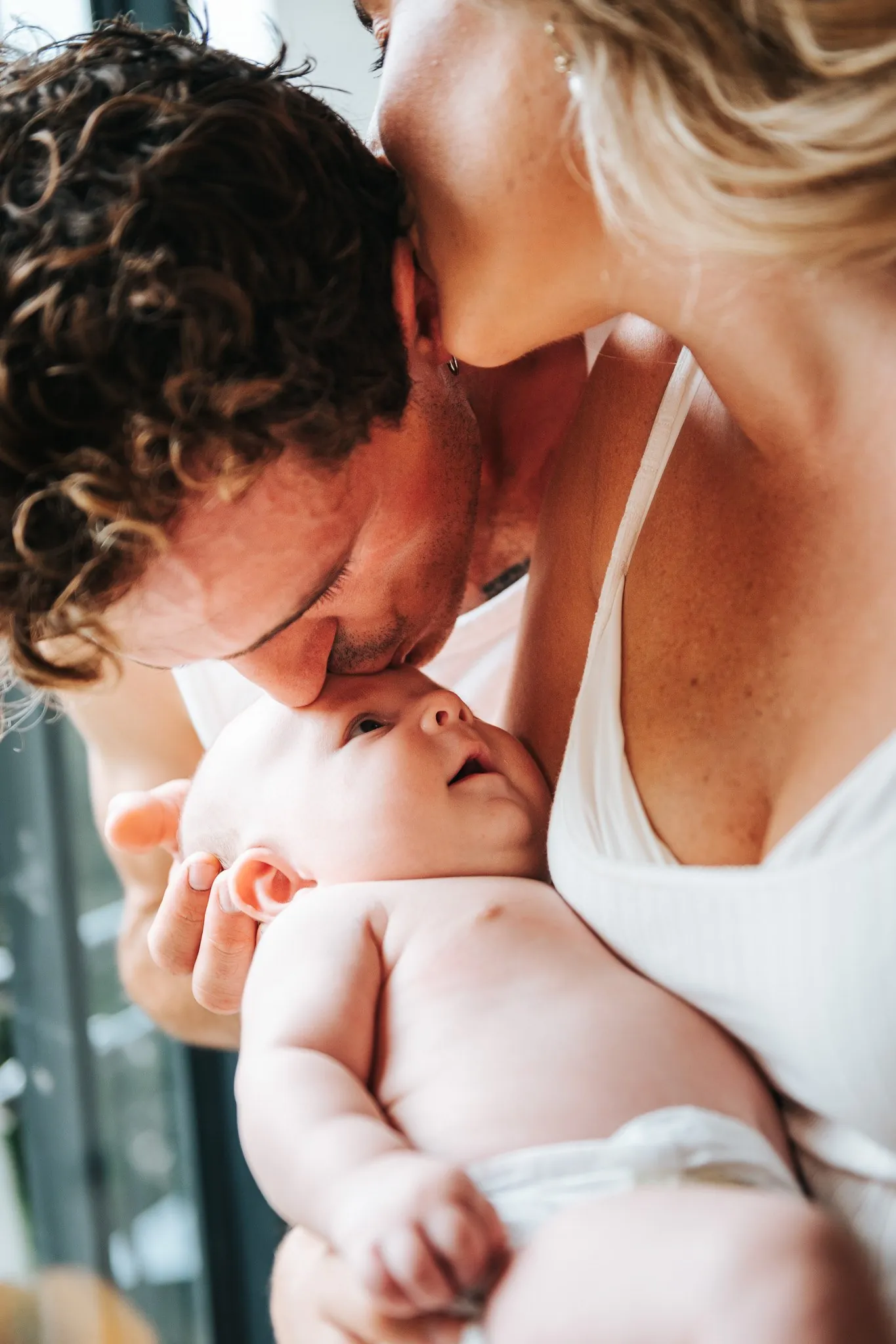 Séance bébé à domicile : papa embrasse le front de son bébé, maman lui embrasse la tête, près d'une fenêtre