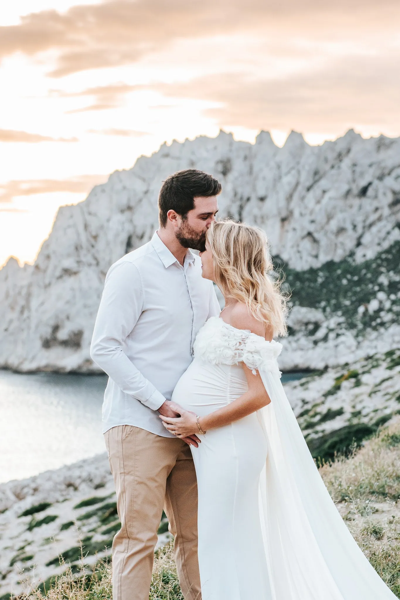 Maternity session in the calanques: a dad-to-be kissing his partner's forehead, she wears a white gown at sunset