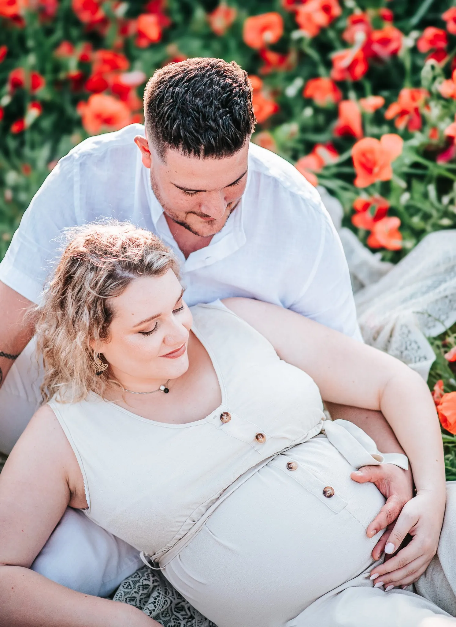 Séance grossesse dans les coquelicots : le couple allongé, main sur le ventre