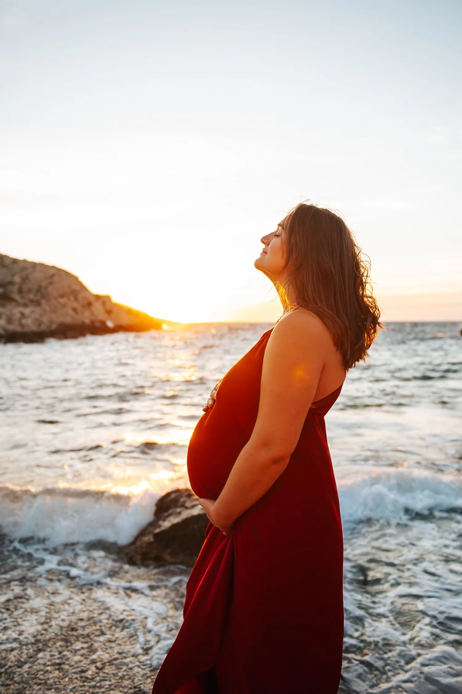 Femme enceinte en robe rouge, yeux fermés et mains sur le ventre, face au soleil couchant au bord de la mer