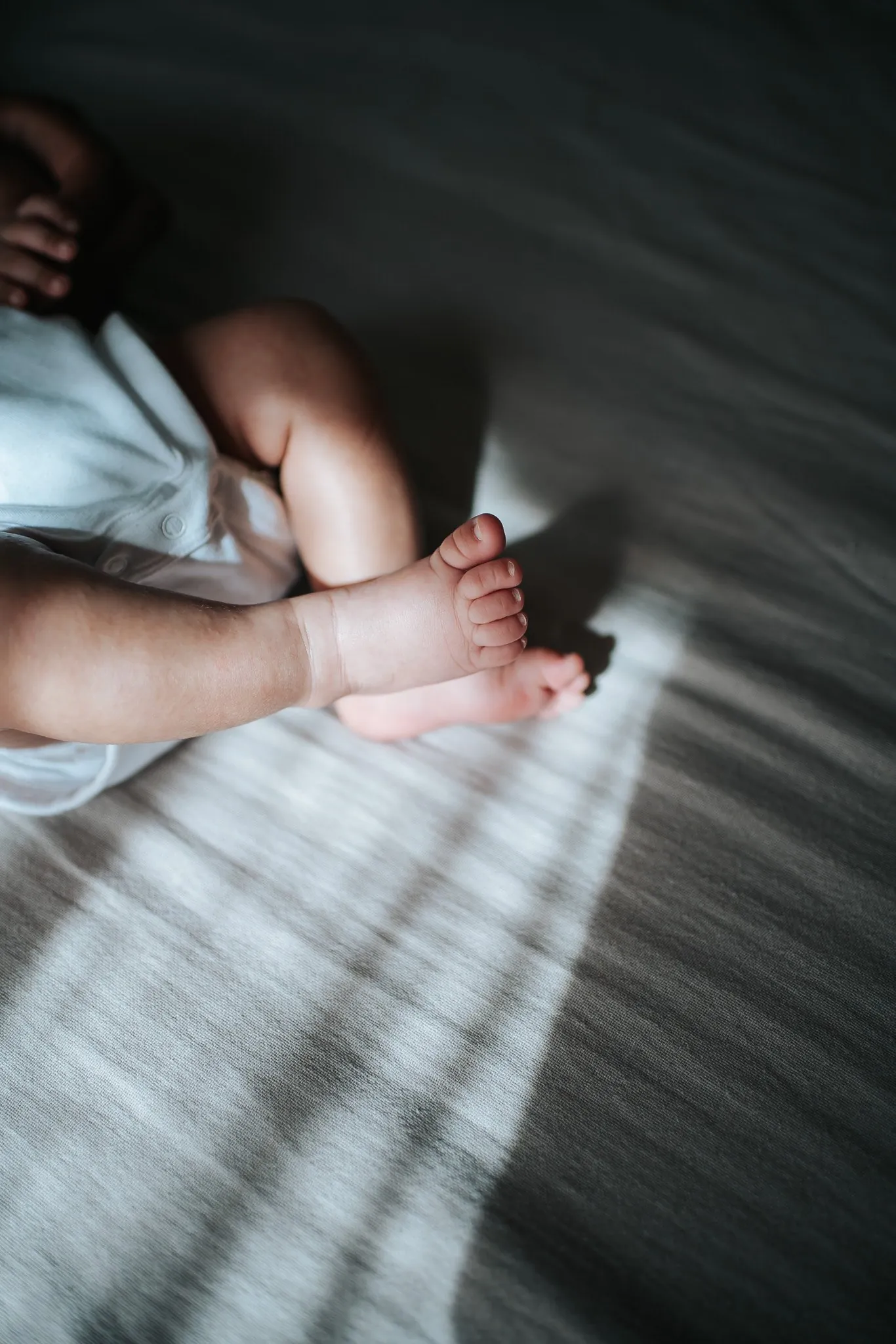 Séance naissance à la maison : jambes et pieds de bébé en body blanc, posés dans un rayon de soleil sur le lit