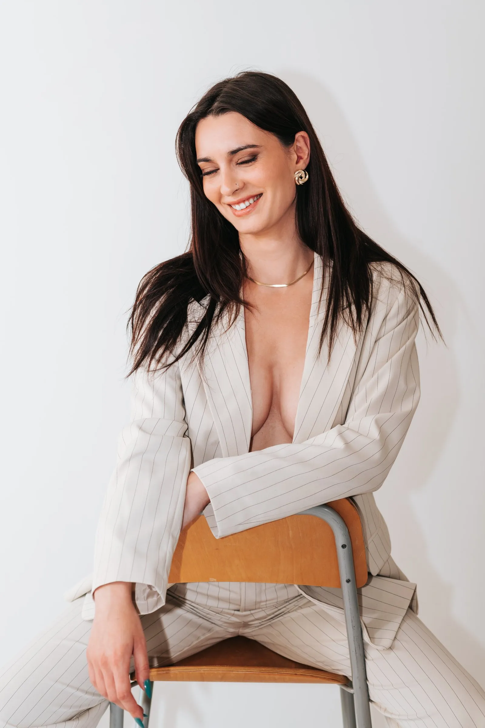 Studio portrait on a white background: a woman in a pinstripe suit sitting backwards on a chair, smiling with eyes closed