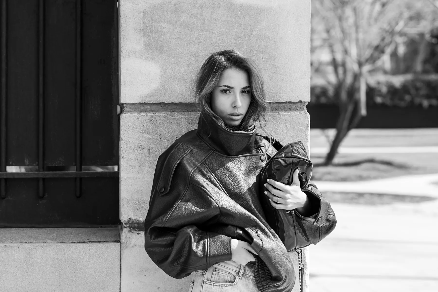 Urban black and white portrait: a young woman in a leather jacket leaning against a stone wall, intense gaze