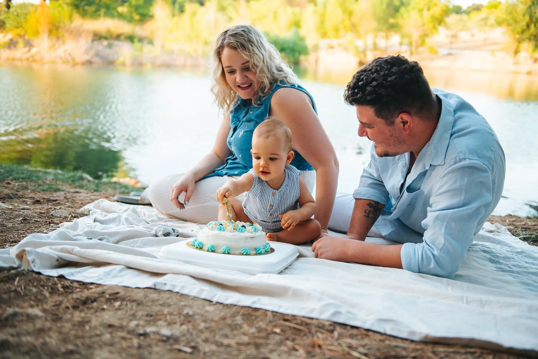 Premier anniversaire au bord d'un lac : bébé qui découvre son gâteau sur un plaid, sous le regard amusé de ses parents