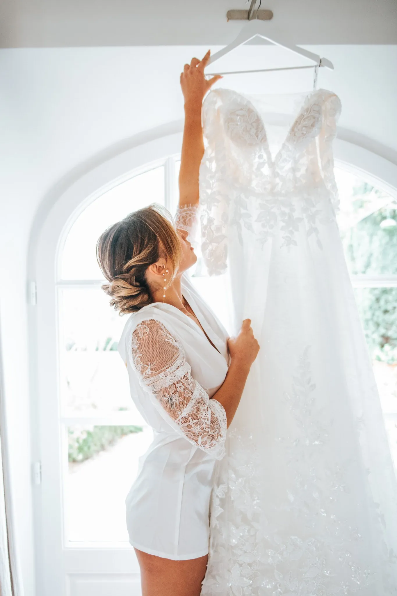 Préparatifs du mariage : future mariée en peignoir de dentelle qui décroche sa robe devant une fenêtre