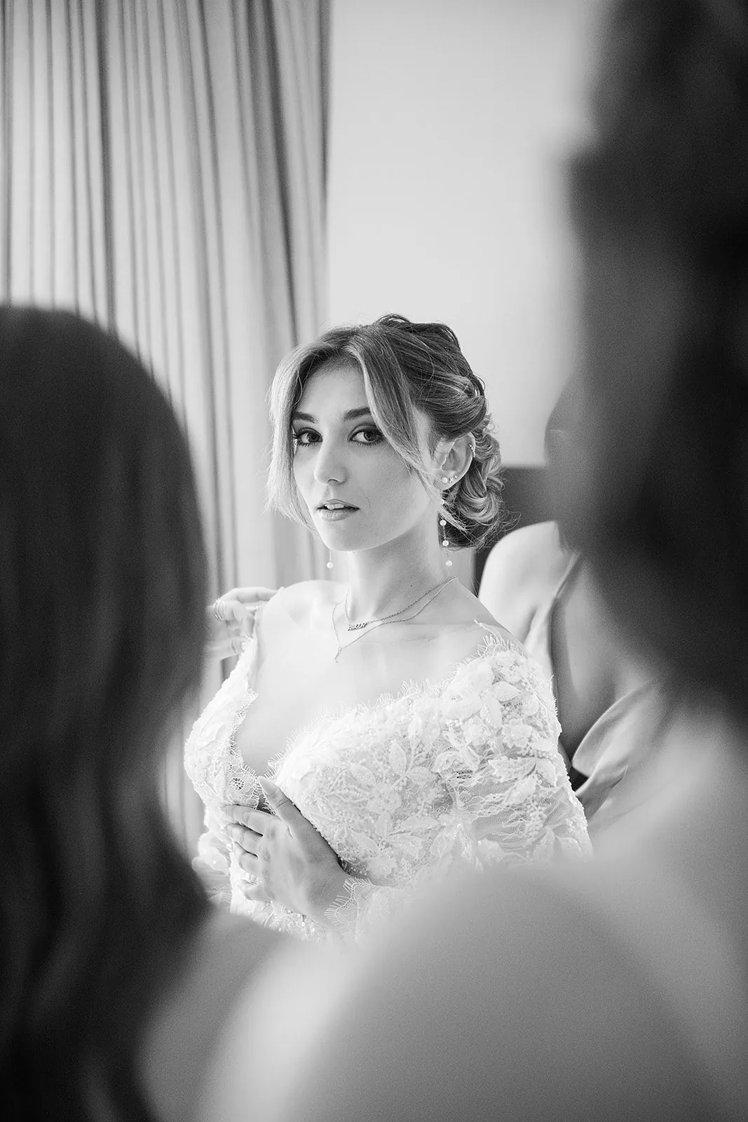 Mariage en noir et blanc : la mariée en robe de dentelle regarde l'objectif pendant qu'on l'aide à s'habiller