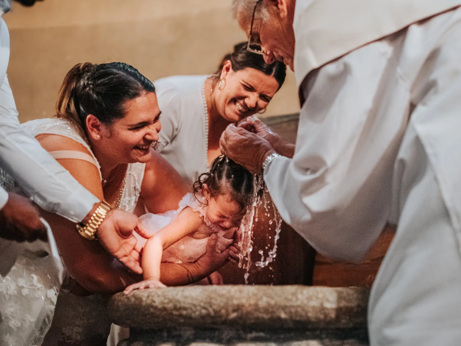 Reportage baptême : le prêtre verse l'eau sur la tête de l'enfant sur les fonts baptismaux, sous les rires de la famille