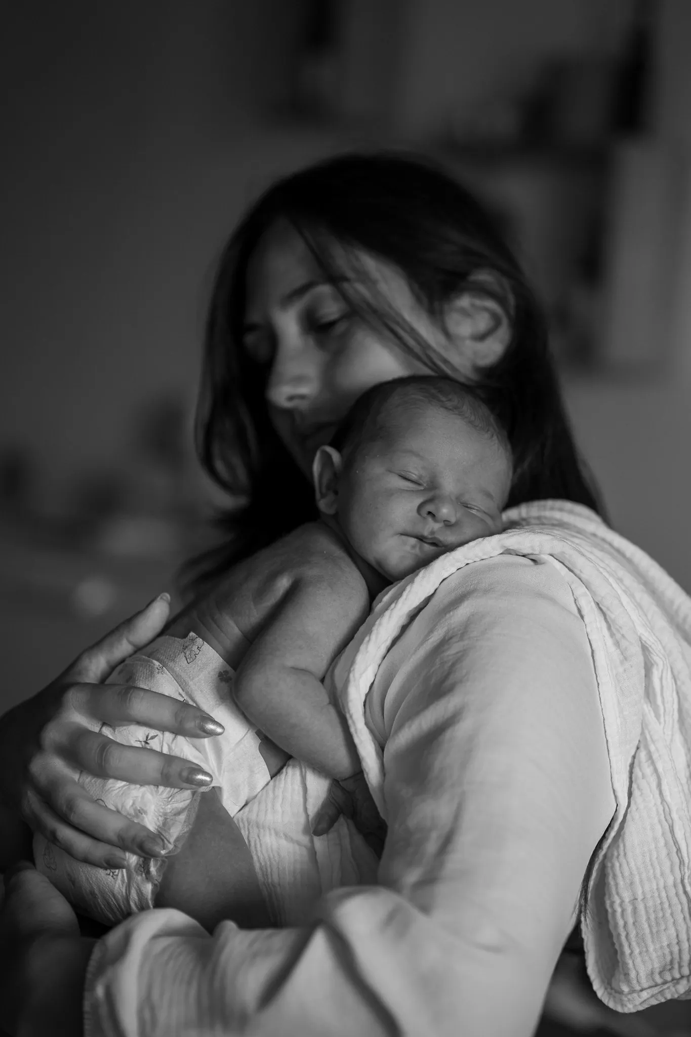 Séance photo bébé à domicile : maman qui serre son nouveau-né endormi contre son épaule, en noir et blanc